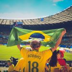 Torcedores brasileiros seguram uma bandeira do Brasil e comemoram nas arquibancadas de um estádio de futebol.