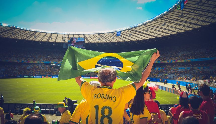 Torcedores brasileiros seguram uma bandeira do Brasil e comemoram nas arquibancadas de um estádio de futebol.