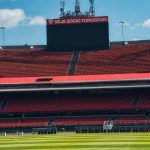 Estádio de futebol vazio, com arquibancadas vermelhas, placar e gramado verde sob céu claro.