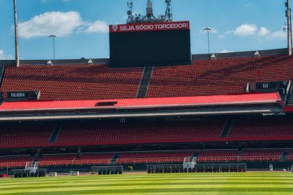 Estádio de futebol vazio, com arquibancadas vermelhas, placar e gramado verde sob céu claro.
