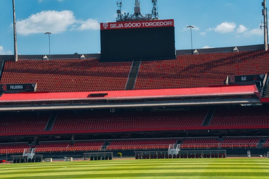 Estádio de futebol vazio, com arquibancadas vermelhas, placar e gramado verde sob céu claro.