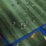 Visão superior de uma partida de futebol em miniatura, com pequenas figuras dispostas sobre um campo.
