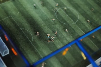 Visão superior de uma partida de futebol em miniatura, com pequenas figuras dispostas sobre um campo.